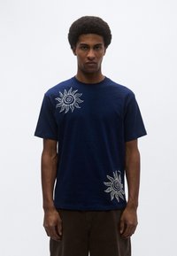 Μπλε ναυτικό βαμβακερό μπλουζάκι με λευκά σχέδια ήλιου στο αριστερό στήθος και στο κάτω δεξί μέρος. Κοντά μανίκια, στρογγυλή λαιμόκοψη και χαλαρή εφαρμογή.