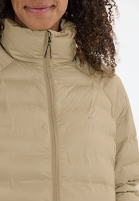 Beige dynejakke med høj krave, lynlås og diskret syning. Har et logo på brystet. Blødt, isoleret materiale.