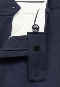 Détail d'une chemise de dress marine montrant la patte de boutonnage avec un bouton noir, un revêtement blanc et des coutures contrastées. Tissu lisse avec une texture subtile.