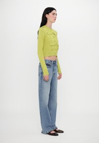 Green cropped cardigan with button front, two chest pockets, and long sleeves, paired with loose-fitting light blue jeans and black flats.