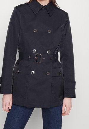 Femme portant un trench-coat noir croisé ceinturé avec des poches boutonnées, sur un jean bleu, sur un fond clair uni.