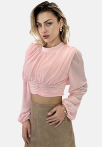 Helles pinkes, gekürztes Blusenshirt mit Falten-Design, langen Ärmeln und gerafftem Taillenbereich; kombiniert mit einem beigen Wildlederrock und einem Armband.