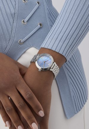 Sudraba rokas pulksteņi ar zilu perlamutra ciparnīcu, romiešu skaitļu atzīmēm un pītā metāla siksniņu, uz struktūras viegli zila auduma fona.