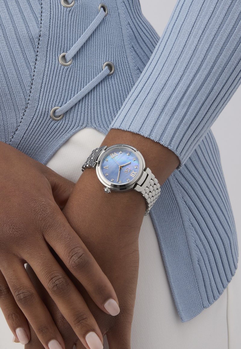 Ceas de mână argintiu cu un cadran perlat albastru, marcaje cu numere romane și o curea din metal împletit, pe un fundal dintr-o țesătură texturată de culoare albastru deschis.