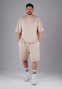 Beige cotton oversized t-shirt with short sleeves, paired with matching shorts. Features logo embroidery. Worn with white sneakers and socks.
