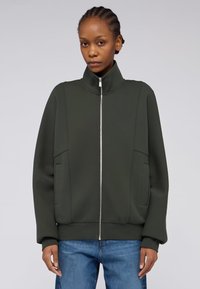 Zelená zipová bunda s vysokým límcem, žebrovanými manžetami a bočními kapsami. Hladká látka s jemnými švy, v kombinaci s modrými džíny.