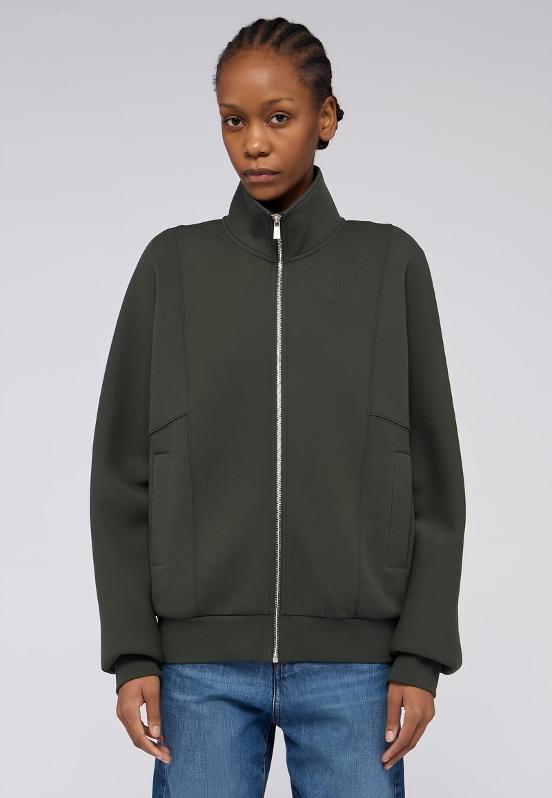Zelená zipová bunda s vysokým límcem, žebrovanými manžetami a bočními kapsami. Hladká látka s jemnými švy, v kombinaci s modrými džíny.