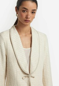 Cremefärgad blazer i strukturerad tyg med sjalkrage, knytning och subtil glans. Slät innerfoder synlig nedanför.