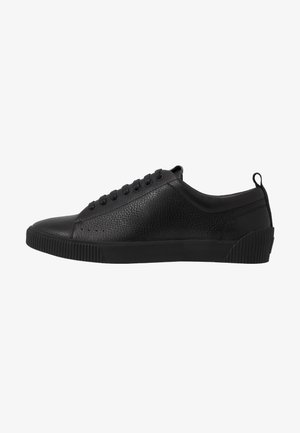 Schwarzer flacher Ledersneaker mit strukturierter Oberfläche, schwarzen Schnürsenkeln, Gummisohle und Fersenlasche, seitlich gezeigt.