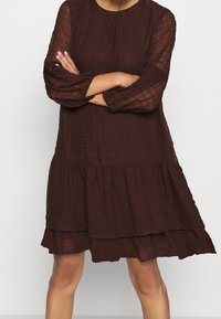 Kvinna stående med armarna i kors, iförd en mörkbrun, texturerad klänning med lång ärm, tierad kjol och genomskinliga ärmar.