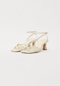 Sandales beige à lanières avec un orteil carré, une bride de cheville ajustable et des détails en doré, dotées d'un talon épais pour plus de stabilité.