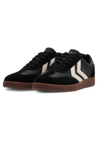 Baskets noires en daim et cuir synthétique, avec des bandes diagonales blanches, une semelle en caoutchouc marron et des accents texturés.