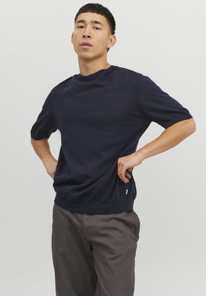 Suéter de punto de manga corta en azul marino con cuello y dobladillo acanalados. Combinado con pantalones gris claro que presentan un detalle de etiqueta lateral.