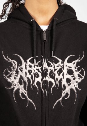 Zwarte zip-up hoodie met abstracte witte grafische tekst op de borst en zwarte trekkoorden met metalen uiteinden.