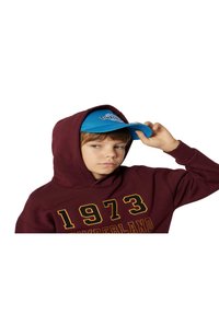Felpa maroon con "1973" in giallo, indossata con un cappellino blu con logo Timbs. Il cappellino ha una visiera curva e pannelli frontali.