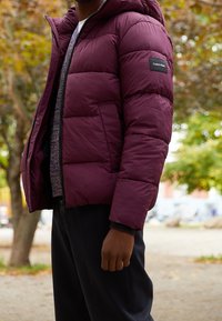 Bordowa kurtka puchowa z kapturem, pikowany wzór, czarna naszywka z logo na górnej części ramienia, noszona na ciemnym swetrze i spodniach, na zewnątrz.