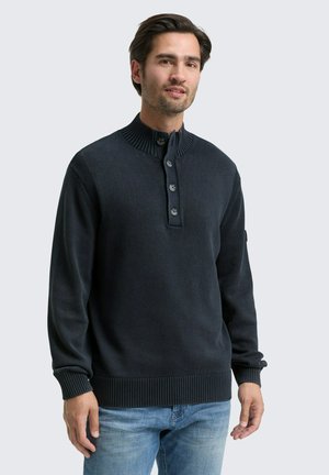 Hombre vistiendo un suéter de punto con cuello abotonado en azul marino oscuro y jeans de mezclilla claros, de pie contra un fondo gris claro liso.