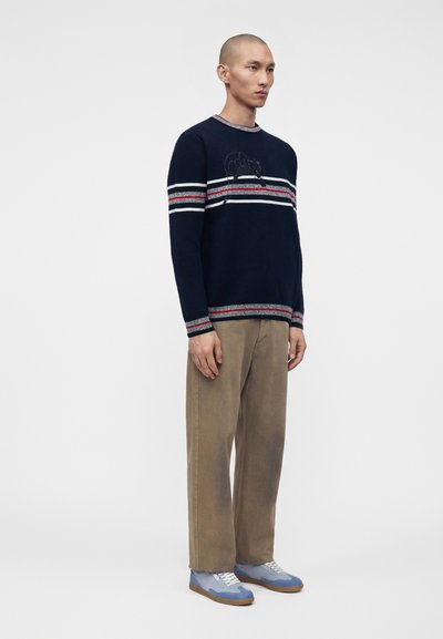 Homme portant un pull marine à rayures rouges et blanches, un pantalon beige à jambes larges et des baskets bleues, debout devant un fond uni.