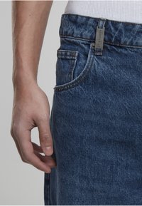 Muška ruka odmara uz džep plavih traperica, bijela rebrasta majica je uvučena unutra.