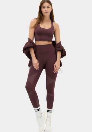 Donna in piedi con reggiseno sportivo bordeaux e leggings con giacca coordinata sulle braccia, calzini bianchi con strisce nere e scarpe da ginnastica bianche.