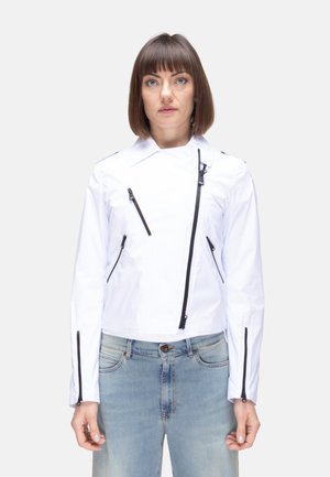 Donna con capelli castano lisci, indossa una giacca bianca con cerniera e jeans azzurri chiari, in piedi davanti a uno sfondo chiaro e semplice.