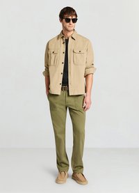 Hellbeiges Hemd mit Knöpfen und zwei Brusttaschen, schwarzes T-Shirt darunter, olivgrüne Hose und beige Schuhe. Schwarze Sonnenbrille vervollständigt den Look.