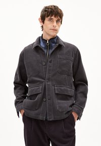 Dunkelgrau Samtjacke mit einem Kragen, Knopfverschluss, zwei vorderen Taschen und strukturierter Stoff. Getragen über einer mit Reißverschluss versehenen Schicht.