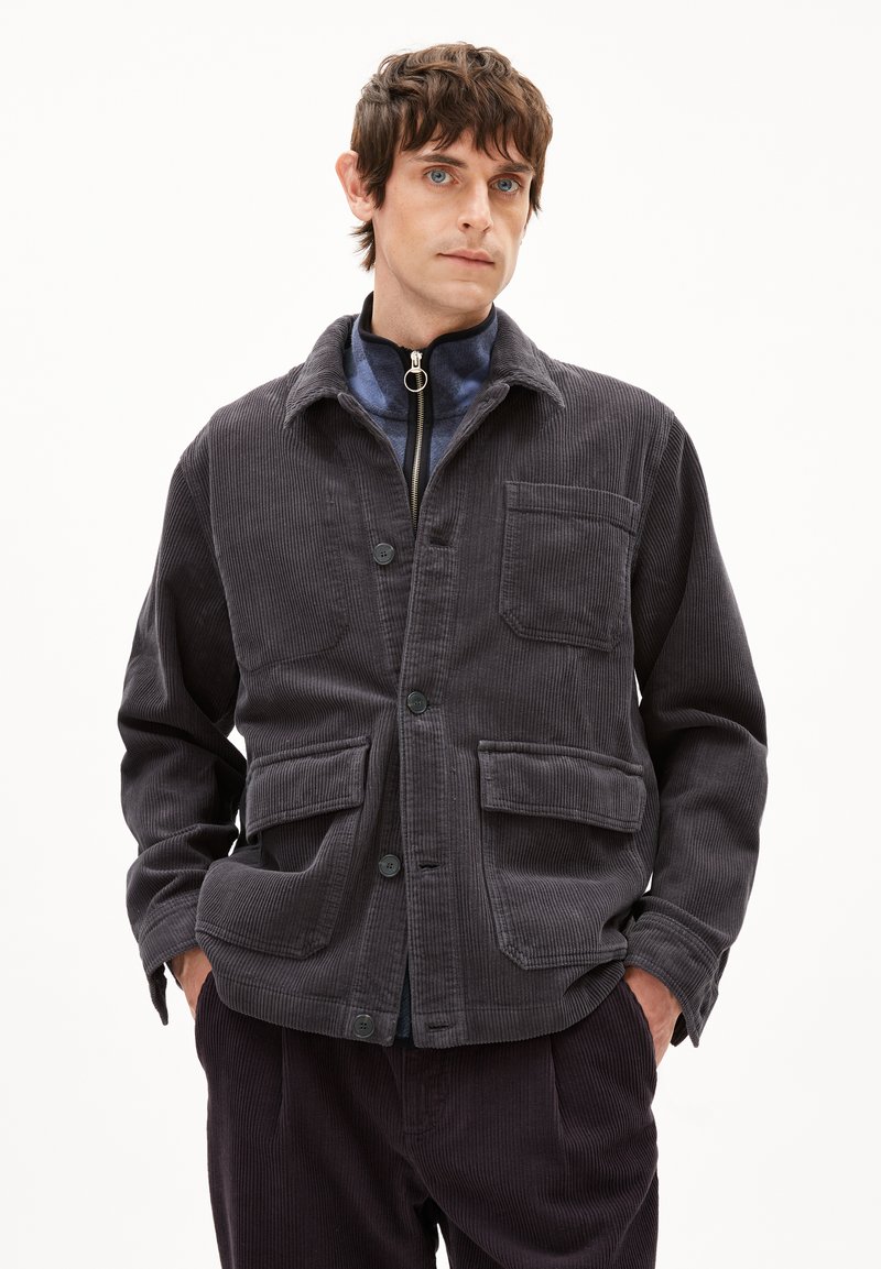 Dunkelgrau Samtjacke mit einem Kragen, Knopfverschluss, zwei vorderen Taschen und strukturierter Stoff. Getragen über einer mit Reißverschluss versehenen Schicht.