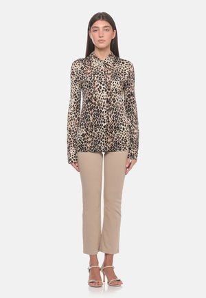 Donna che indossa una camicetta a maniche lunghe con stampa leopardata, pantaloni beige corti e sandali beige con tacco alto, in piedi davanti a uno sfondo semplice.