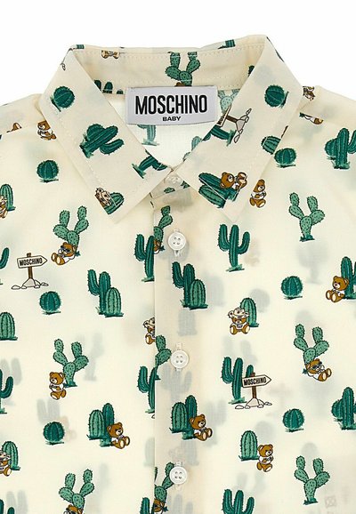 Maglietta a maniche corte per bambini di colore beige chiaro con stampa di cactus verdi e orsetto. Presenta un colletto classico e bottoni bianchi. Etichetta del marchio: Moschino Baby.