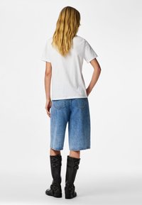 Šviesiaplaukis asmuo stovi nugara, dėvi paprastą baltą marškinėlį, mėlynos spalvos džinsinius trumpus kelnes iki kelių ir juodus kelių ilgio batus su sagtimis.