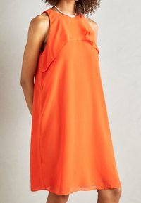 Robe orange sans manches en tissu léger, avec un design à volants au niveau de l'encolure et une coupe ample et décontractée.