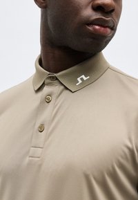 Άνδρας που φοράει μπεζ πόλο με τρία κουμπιά και μικρό λευκό κεντημένο λογότυπο στο γιακά.