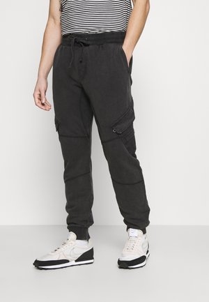 Mężczyzna w czarnych joggerach typu cargo z elastycznym pasem i białych i czarnych tenisówkach, stojący na białym tle.