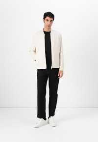 Light beige bomber jacket with a front zipper, paired with a black t-shirt and black trousers. Model wears white sneakers, and stands against a plain background.