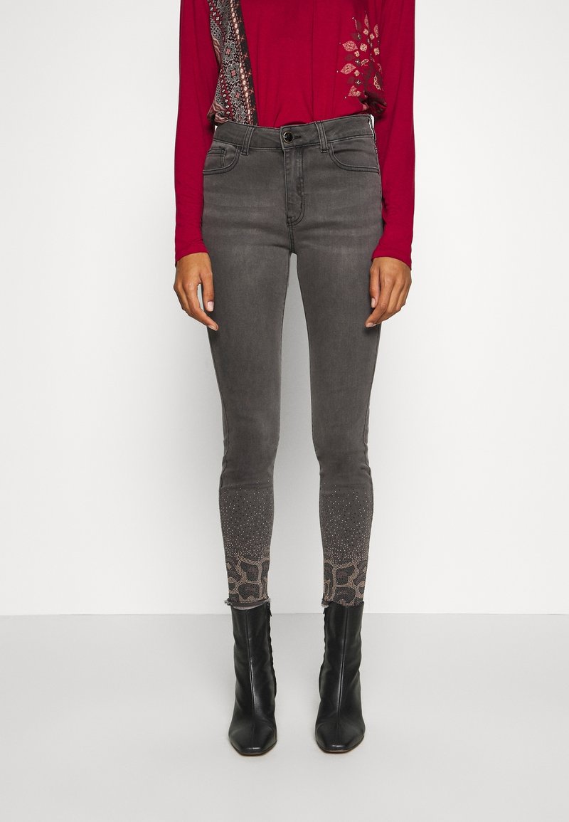 Femme portant un jean skinny gris avec des ourlets cloutés et à motifs, un chemisier rouge à manches longues et des bottines noires, sur un fond uni.