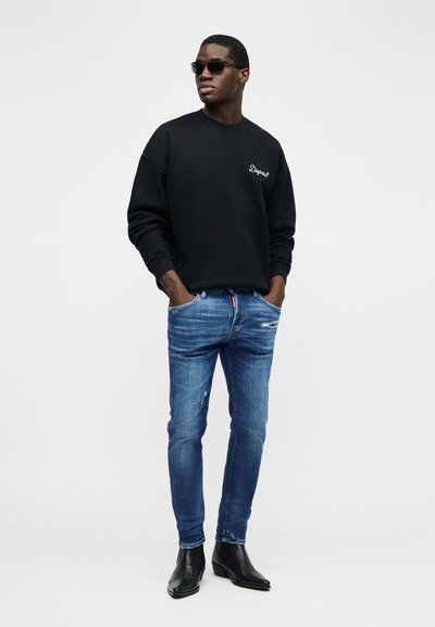 Άνδρας που φοράει μαύρο φούτερ, μπλε σκισμένο στενό τζιν, μαύρα μποτάκια και γυαλιά ηλίου, στέκεται με τα χέρια στις τσέπες.