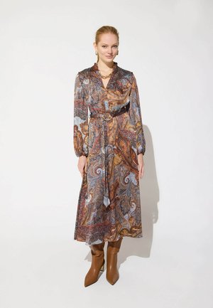 Femme portant une robe à manches longues ceinturée avec un motif paisley marron, bleu et orange, assortie à des bottines marron et des bijoux dorés.