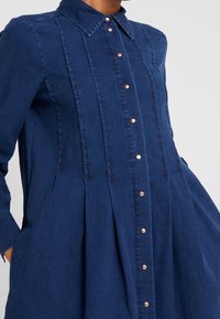 Džínové šaty v tmavě modré barvě, s knoflíkovým zapínáním na přední straně, růžově zlatým kováním, límečkem a plisovanou sukní.
