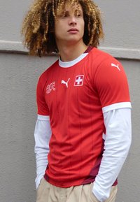 Rød atletisk trøje med hvide lange ærmer, der har et schweizisk flaglogo og Puma-branding. Tekstureret stof med lodrette striber.