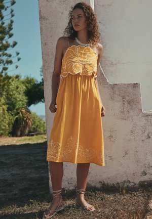 Young woman in a yellow sundress with white floral embroidery and sandals standing outdoors beside a weathered white wall under clear sky.