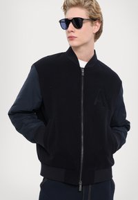 Giacca bomber navale con corpo in lana e maniche in nylon, dotata di chiusura con zip e tasche frontali. Logo ricamato sul petto.