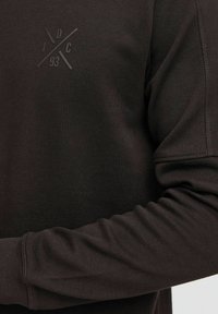 Dark brown long-sleeve top featuring textured fabric, ribbed cuffs, and a subtle embossed logo in the upper left corner.