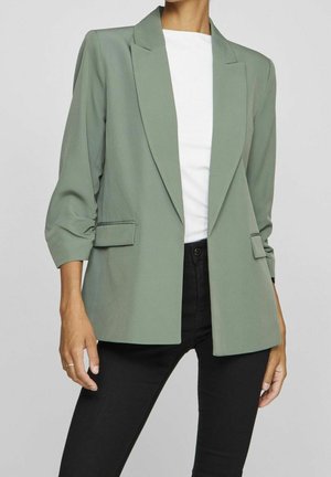 Femme portant un blazer vert sauge à manches trois-quarts, un top blanc et un pantalon noir, debout devant un fond gris uni.