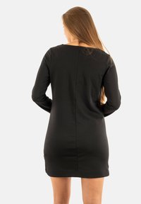 Frau mit langen braunen Haaren, die ein enganliegendes schwarzes Langarmkleid trägt, mit den Händen in die Hüften gestützt, stehend und sich von einem schlichten weißen Hintergrund abwendend.