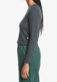 Graues Langarmshirt mit einer taillierten Passform, gerippter Textur und cropped Länge; getragen mit grünen Cordhosen mit Seitentaschen.