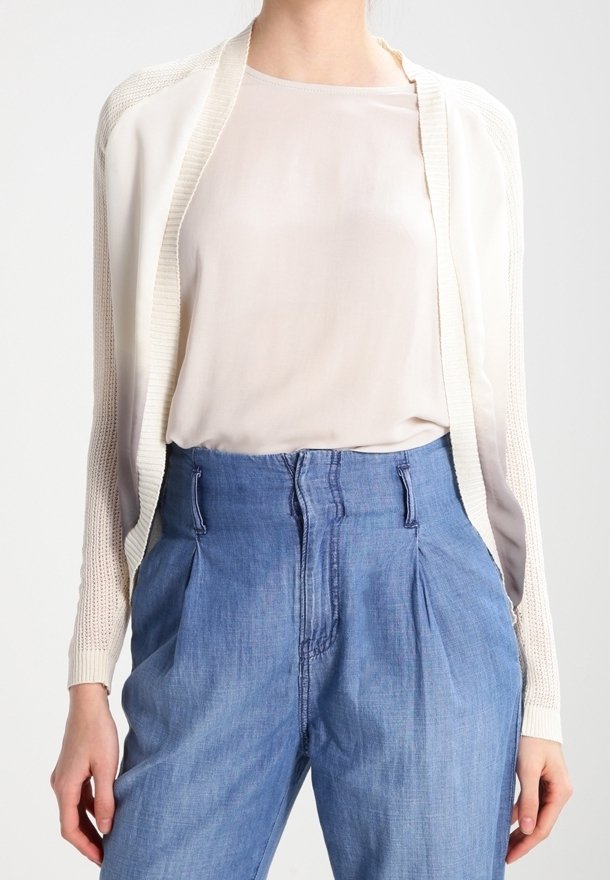 Femme portant un chemisier blanc transparent, un cardigan crème tricoté et un pantalon en jean bleu taille haute avec des plis et une fermeture éclair sur le devant.