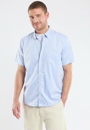 Chemise à boutons à manches courtes bleu clair avec un col étalé et des boutons en tissu, présentant une texture subtile et des coutures latérales pour un ajustement ajusté.