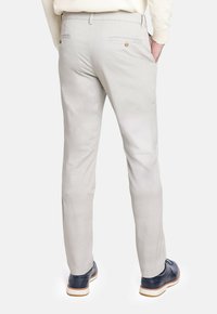 Hombre vestido con pantalones slim-fit beige claros con bolsillos traseros con botón y zapatos de vestir azul marino, de pie con una mano en el bolsillo.