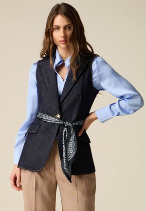 Donna con lunghi capelli castani che indossa una camicia azzurro chiaro, un gilet smanicato in denim scuro con cintura a sciarpa fantasia e pantaloni beige.